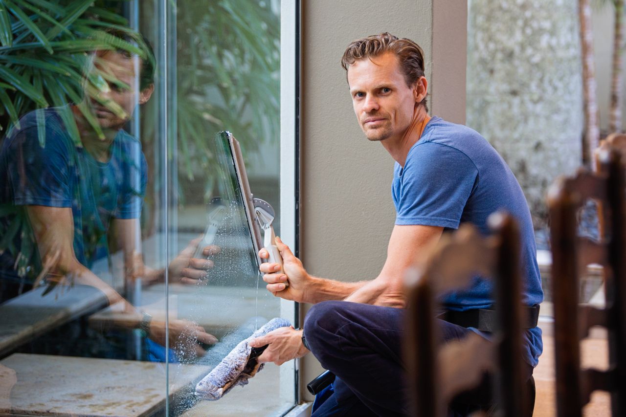project-01-d A man in a blue t-shirt cleaning large glass pane with a squeegee indoors.