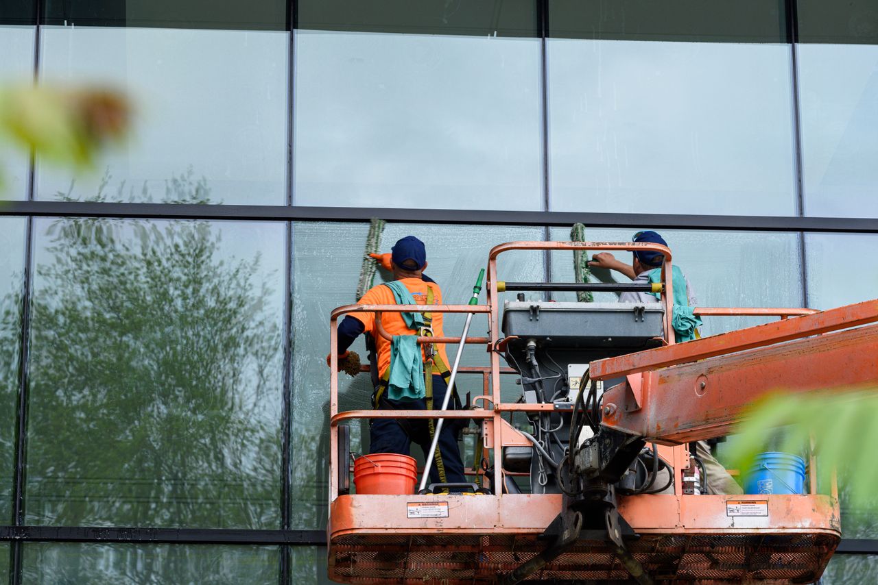 services-01 Deux travailleurs nettoient les fenêtres d'une façade en verre à l'aide d'une nacelle élévatrice, ce qui améliore l'esthétique du bâtiment.